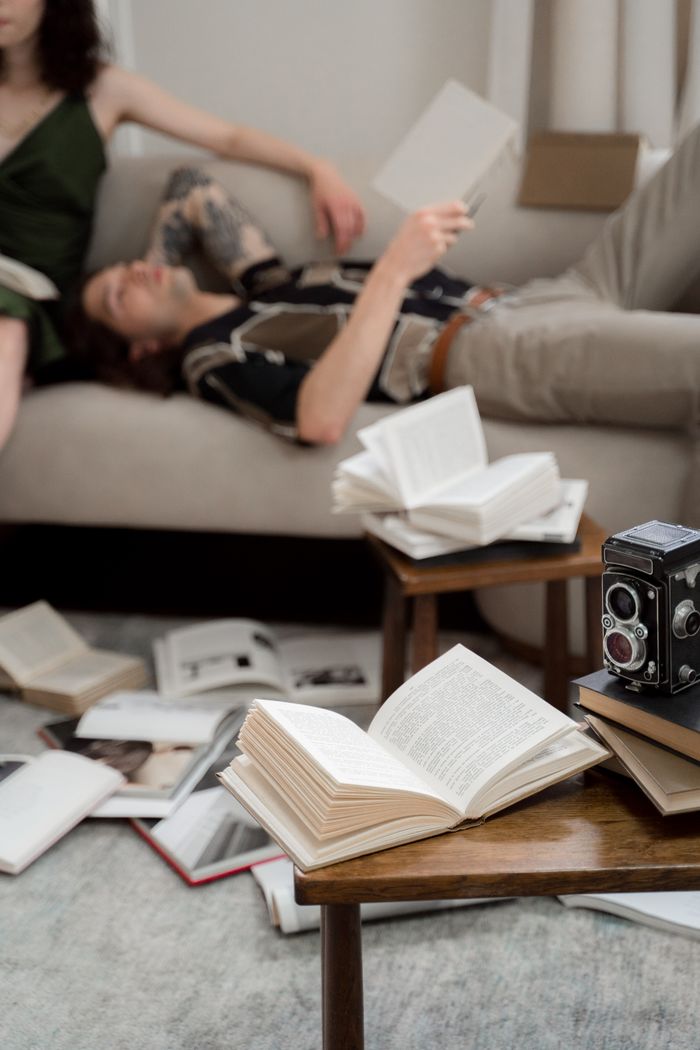 Young couple relaxing and reading in a cluttered room with open books and a vintage camera nearby.